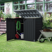 Load image into Gallery viewer, The Keter Hideaway Storage Shed XL Grey outside in the garden. The box has its lid open and a spade, watering can and hose pip stored inside. 
