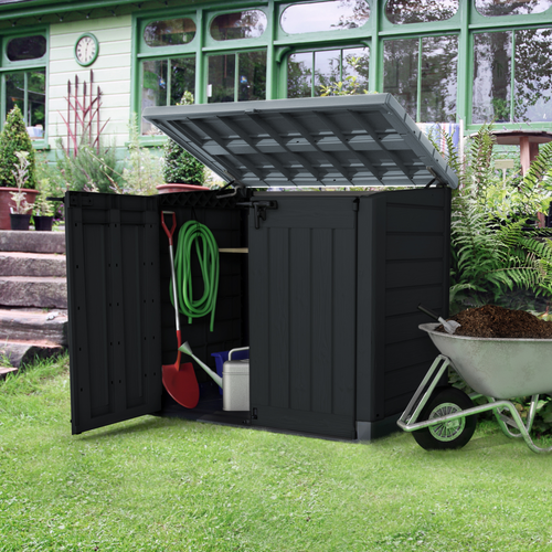 The Keter Hideaway Storage Shed XL Grey outside in the garden. The box has its lid open and a spade, watering can and hose pip stored inside. 