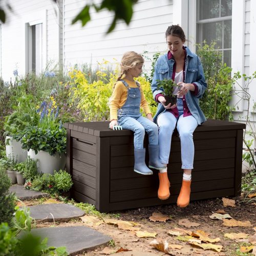 The Keter Hingham dark brown storage box outside in the garden with a child and a woman using it as a bench. 