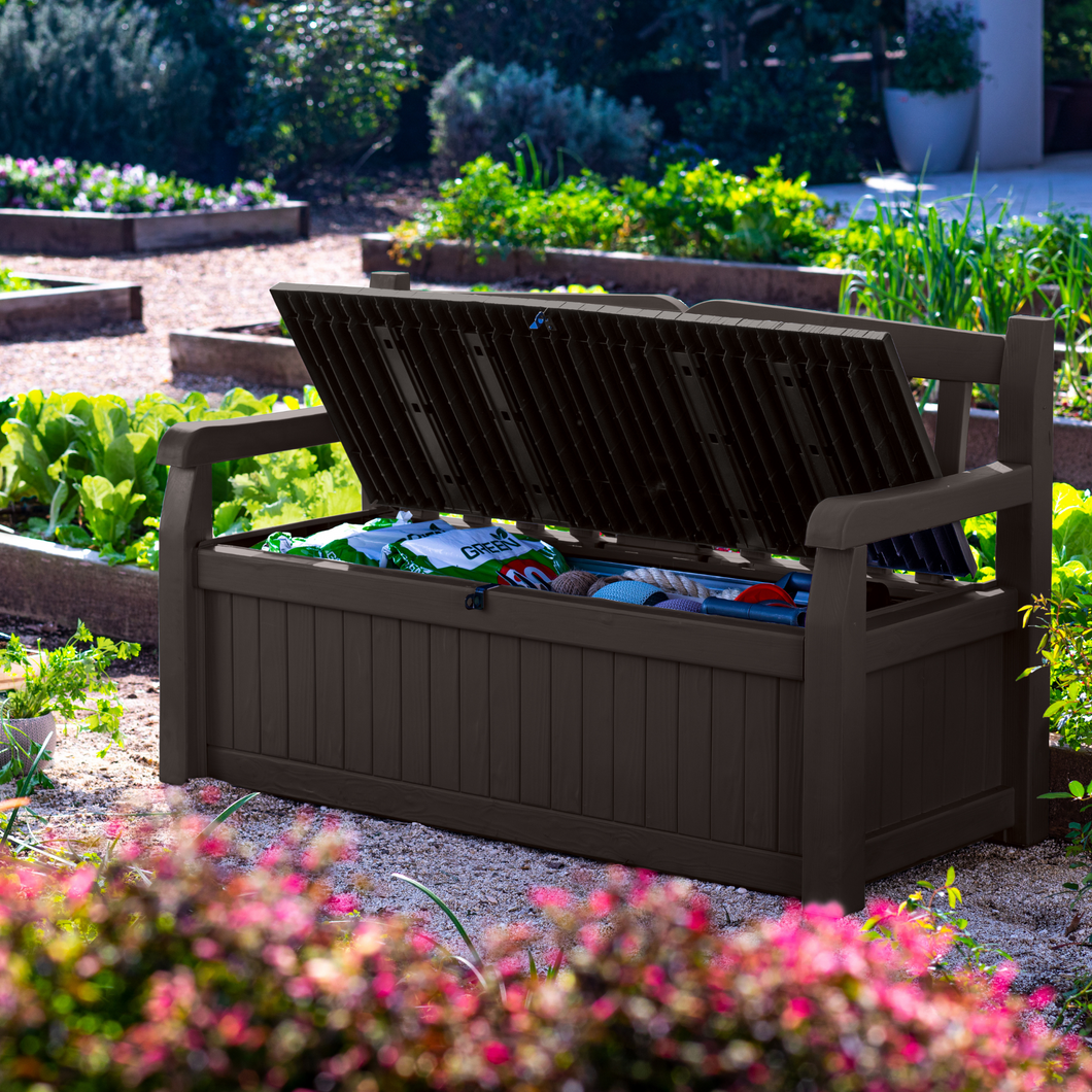 The Keter Iceni Storage Bench 265L Brown outside in the garden. The bench is open and shows garden accessories inside. 