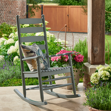 Load image into Gallery viewer, The Oakwell grey rocking chair in the garden with a pillow and flowers behind in the background. 
