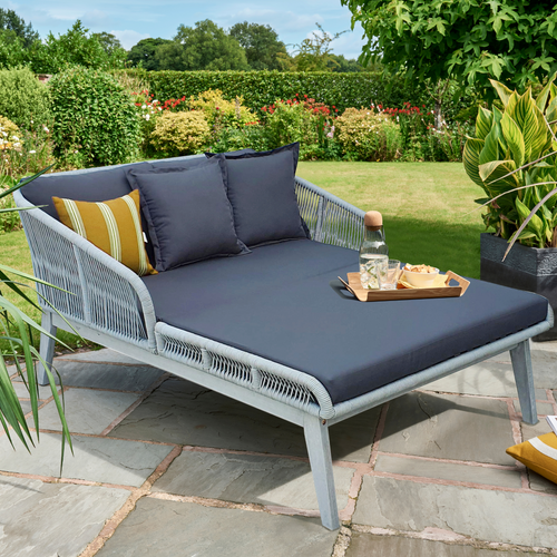 The Dara Day Bed in Grey Rope and Greywash outside in the garden. The daybed has scatter cushions and a tray placed on it. 