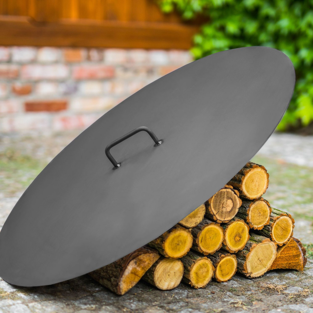 The Cook King Steel Lid for 80cm fire pits leant against some wooden logs outside. 