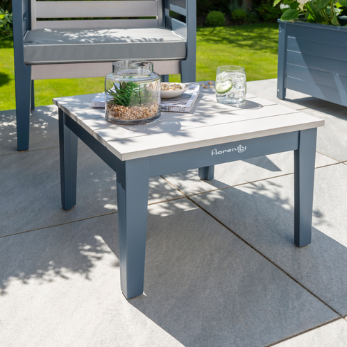 The Florenity Galaxy Side Table on the garden patio with a drink and magazine placed on the table top. The Florenity Galaxy chair is shown in the background. 