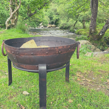 Load image into Gallery viewer, The 60cm fire pit at the side of a river on the grass bank. The fire pit is stood on its stand with firewood inside. 
