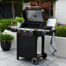Load image into Gallery viewer, The Norfolk Grills Atlas 300 with Side Burner outside in the garden. The side burner has a pan on, hanging tea towel and various condiments in the side shelf. 
