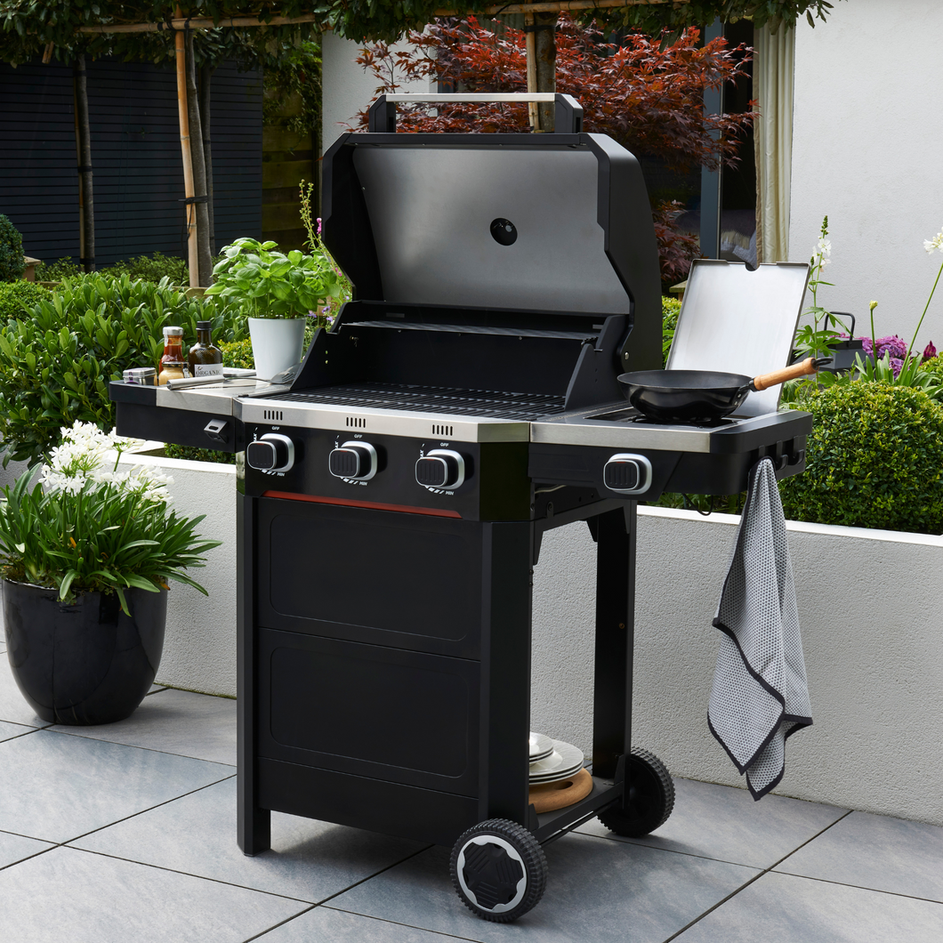 The Norfolk Grills Atlas 300 with Side Burner outside in the garden. The side burner has a pan on, hanging tea towel and various condiments in the side shelf. 