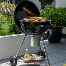 Load image into Gallery viewer, The Corus charcoal bbq on a garden patio with a variety of food cooking on the grill. There are green plants in the background and a tray with lemons and condiments on the bbq storage shelf which sits under the bbq. 
