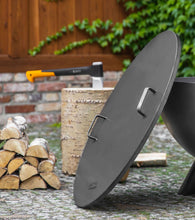 Load image into Gallery viewer, The Cook King fire pit lid with two handles leant against a fire pit with wood logs in the background.

