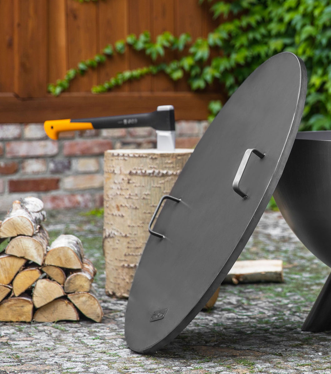 The Cook King fire pit lid with two handles leant against a fire pit with wood logs in the background.