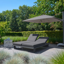 Load image into Gallery viewer, The Fitz Roy double sunlounger outside in the garden. The parasol gives the double lounger shade and there is a table with some accessories on it. 

