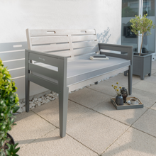 Load image into Gallery viewer, The Florenity Grigio Two Seat Bench Set outdoors against a white wall and plants either side. There is a tray below the bench with a cup of coffee and a plant on. 
