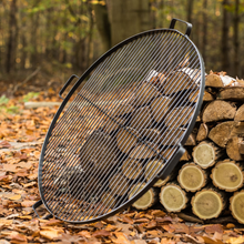 Load image into Gallery viewer, Cook King natural steel grill with 4 handles leant against a wood pile in the forest. The ground covered with leaves and tree trunks in the background. 
