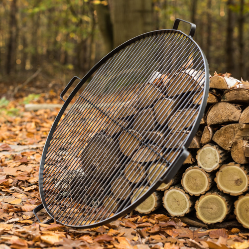 Cook King natural steel grill with 4 handles leant against a wood pile in the forest. The ground covered with leaves and tree trunks in the background. 