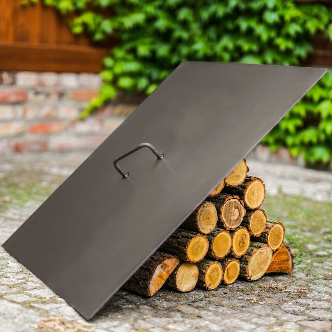 Cook king 70cm square fire pit lid leant against a pile of wooden logs in a garden. 