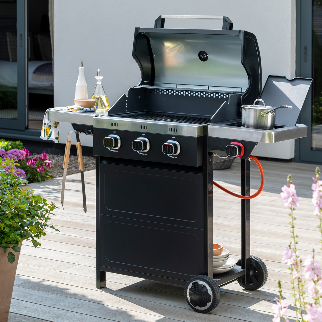 The Norfolk grills vista 300 gas BBQ grill on the garden decking outside with the lid open. The bbq has various condiments and accessories on it. 